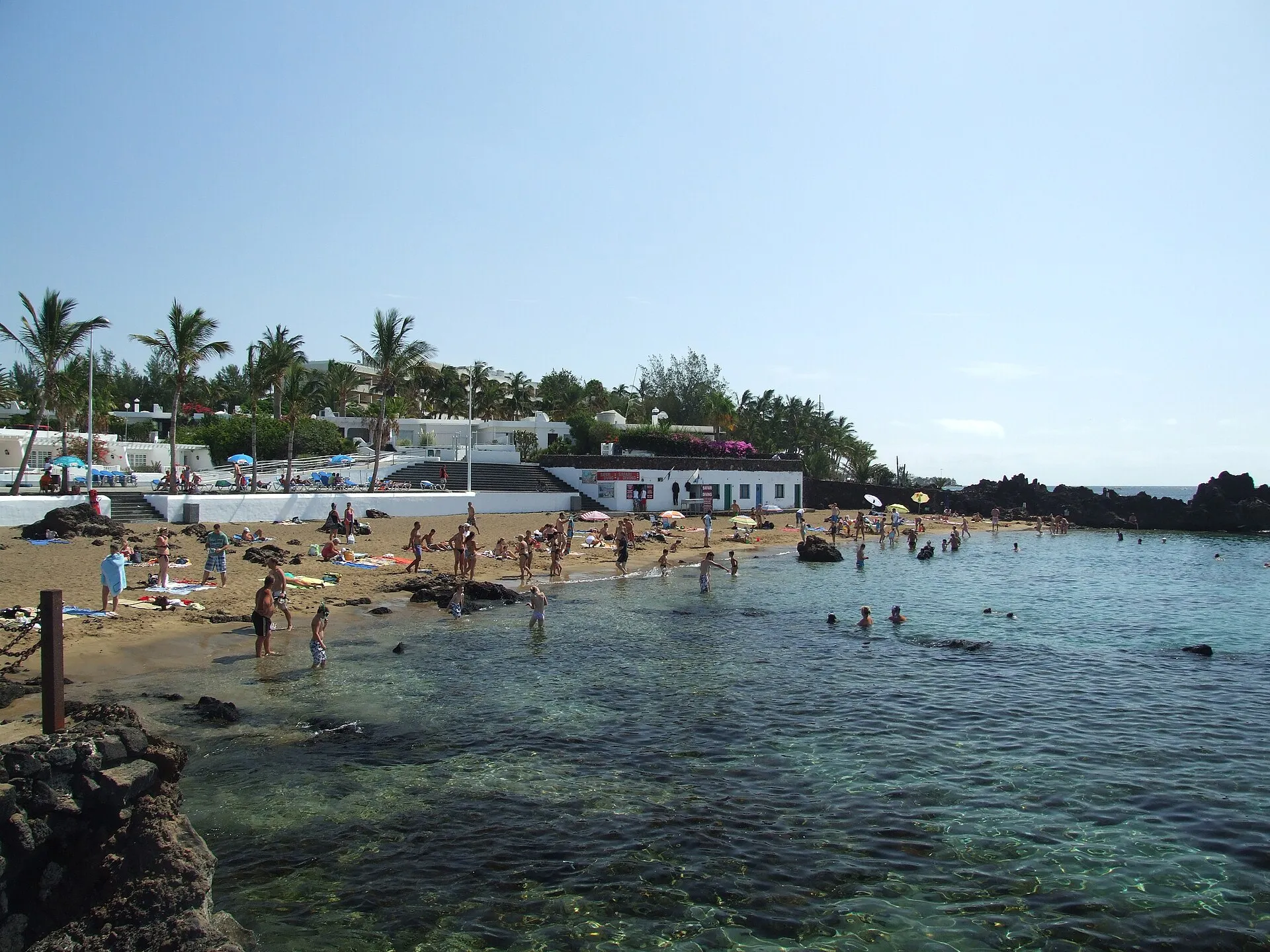 Playa Chica: a small sandy cove between black lava rocks, with clear shallow water and bathers